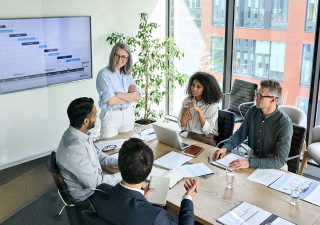 Senior female ceo and multicultural business people discussing company presentation at boardroom table. Diverse corporate team working together in modern meeting room office. Top view through glass